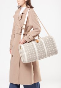 Borsa a tracolla bianca decorata con un motivo geometrico beige, hardware dorato e una tracolla rimovibile, tenuta da una persona in un trench beige.