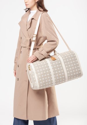 Sac de voyage blanc à motifs avec un design géométrique beige, accessoires en or et une bandoulière amovible, tenu par une personne portant un trench-coat beige.