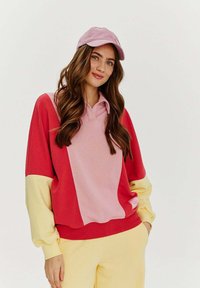 Mikina s blokováním barev, která obsahuje růžové, červené a žluté části, vyrobená z měkké tkaniny s vystřiženým límcem. K outfitu se skvěle hodí odpovídající růžová čepice.