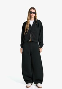 Zwart oversize sweatshirt met rits en voorzak, gecombineerd met bijpassende baggy broek. Gedragen met witte sneakers en zwarte zonnebril.