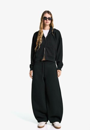 Zwart oversize sweatshirt met rits en voorzak, gecombineerd met bijpassende baggy broek. Gedragen met witte sneakers en zwarte zonnebril.