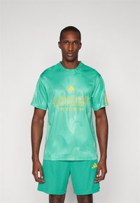 Top de desporto em malha verde com o logotipo amarelo "ADIDAS", apresentando um pescoço redondo e mangas curtas. Calções verdes a condizer em baixo.