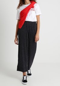 Bílý triko s červenou grafikou, doplněné černými pruhovanými širokými kalhotami a černými teniskami. Červený strukturovaný svetr přehozený přes rameno.