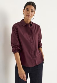 Bordeaux overhemd met knopen, voorzien van een klassieke kraag, lange mouwen met elastische manchetten en een gladde, gestructureerde stof, gestyled met donkere jeans.