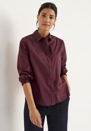 Bordeaux overhemd met knopen, voorzien van een klassieke kraag, lange mouwen met elastische manchetten en een gladde, gestructureerde stof, gestyled met donkere jeans.