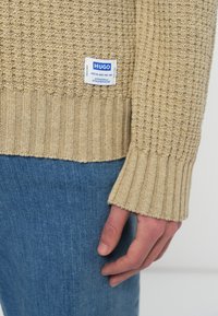 Beżowy sweter z dzianiny z prążkowanym dołem i mankietami, z niebieską metką z napisem "HUGO." Na drugim planie jeansy.