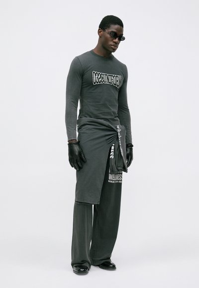 Mann mit dunkler Sonnenbrille, einem enganliegenden Langarmhemd mit Aufdruck, weiten Hosen, Handschuhen und einem um die Taille gebundenen, verknoteten Langhemd.