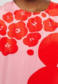Top con estampado floral que presenta grandes flores rojas sobre un fondo rosa, con una textura suave y lisa y un diseño de escote redondo.