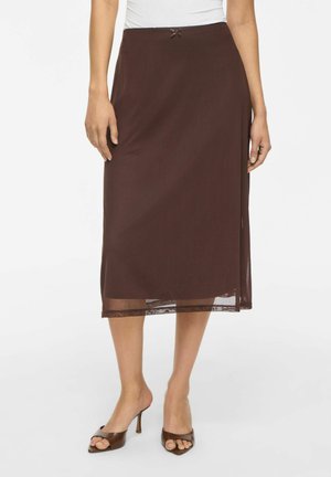 Femme portant une jupe midi marron foncé avec un petit nœud à la taille et des sandales à talons ouvertes marron, debout sur fond blanc.