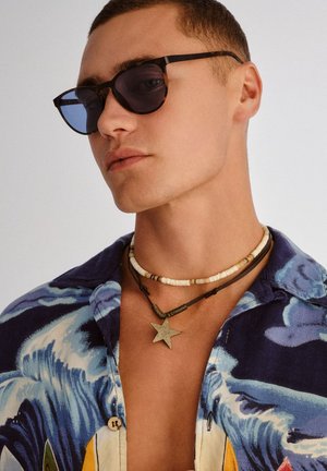 Młody mężczyzna w ciemnych okularach przeciwsłonecznych, niebieskiej, wzorzystej koszuli oraz dwóch naszyjnikach — jednym z koralików, drugim z zawieszką w kształcie gwiazdy.