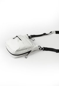 Borsa a spalla in tessuto bianco con tracolla nera regolabile, compartimento singolo con zip e tasca frontale. Presenta un design piatto e una marca discreta.