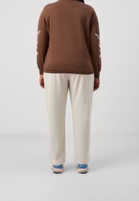 Felpa marrone con accenti decorativi bianchi sulle maniche, abbinata a pantaloni beige chiaro. Le calzature presentano una combinazione di tonalità blu e rosa.