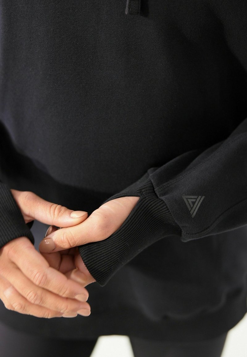 Personne portant un sweat-shirt noir à manches longues avec des orifices pour le pouce, mains doucement jointes devant, logo à motif sur le bord de la manche gauche.