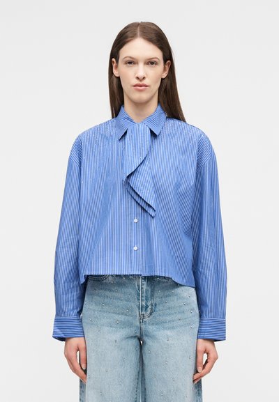 Femme portant un chemisier à fines rayures bleu avec un volant à l'avant et un jean bleu clair orné de petites clous.