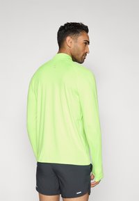 Αθλητικό πουκάμισο με μακριά μανίκια σε έντονο πράσινο λάιμ, κατασκευασμένο από ελαφρύ ύφασμα· διαθέτει ψηλό γιακά και διακριτικές λεπτομέρειες στην πλάτη· συνδυάζεται με σκούρα σορτς.