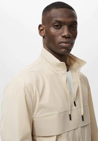 Beige zip-jakke med høj krave, der har en frontlomme, teksturerede paneler og kontrasterende sorte snore. Glat stoftekstur.
