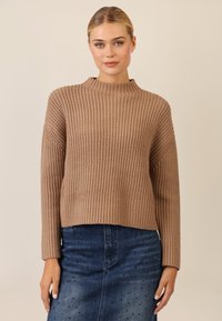 Beige gestrickter Pullover mit hohem Kragen, geripptem Textur und langen Ärmeln, kombiniert mit einem blauen Jeansrock mit dekorativen Akzenten.