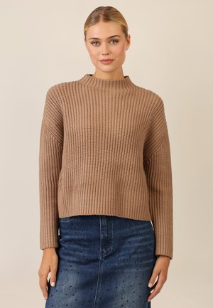 Beige gestrickter Pullover mit hohem Kragen, geripptem Textur und langen Ärmeln, kombiniert mit einem blauen Jeansrock mit dekorativen Akzenten.