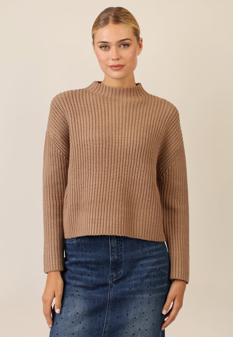 Beige gestrickter Pullover mit hohem Kragen, geripptem Textur und langen Ärmeln, kombiniert mit einem blauen Jeansrock mit dekorativen Akzenten.
