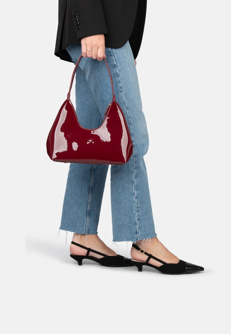 Borsa in pelle verniciata bordeaux a forma di hobo, superficie liscia e manico singolo, tenuta da una persona che indossa jeans azzurri chiari e scarpe nere.