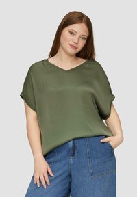 Oliwkowa, luźno dopasowana bluzka z dekoltem w serek i podwini�ętymi rękawami, wykonana z gładkiego materiału. W zestawie z niebieskimi spodniami dżinsowymi o szerokich nogawkach.