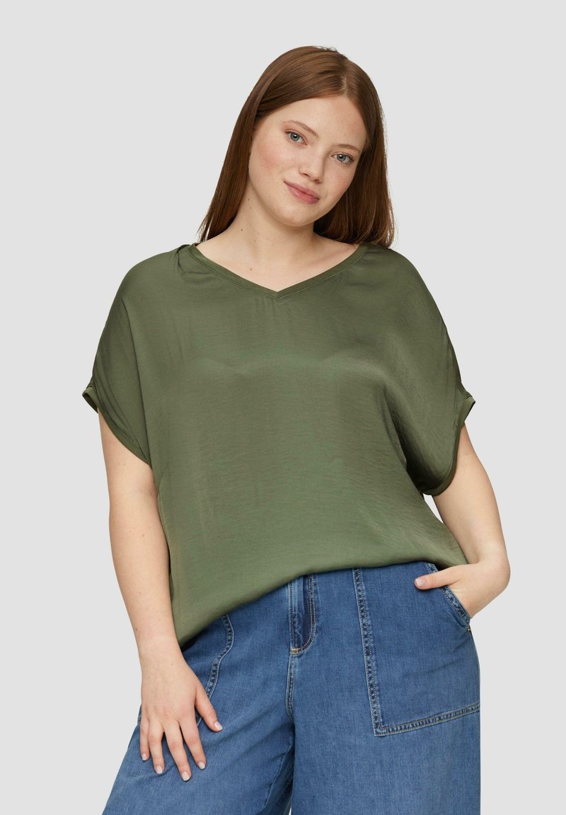Oliwkowa, luźno dopasowana bluzka z dekoltem w serek i podwiniętymi rękawami, wykonana z gładkiego materiału. W zestawie z niebieskimi spodniami dżinsowymi o szerokich nogawkach.
