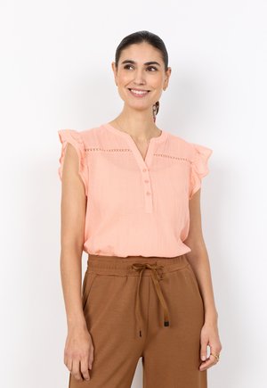 Lächelnde Frau mit dunklen Haaren in einer hellpfirsichfarbenen, ärmellosen Bluse mit Rüschen und braunen Hosen mit Kordelzug, vor weißem Hintergrund stehend.