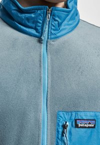 Giacca in fleece blu con colletto alto, zip azzurra chiara e logo Patagonia su una tasca a zip sul petto.