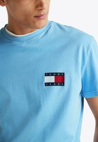 Camiseta de algodón azul claro con un pequeño logo rectangular de Tommy Jeans en azul marino, blanco y rojo. Cuello redondo estándar y mangas cortas.