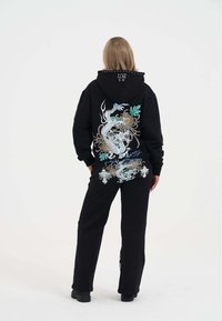 Zwarte hoodie en broekset met kleurrijke draak borduursel en bloemmotieven op de achterkant, gemaakt van een zachte stof met een ontspannen pasvorm.