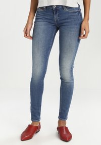 Vaqueros skinny de mezclilla azul con un diseño desgastado, que presentan una cintura de altura media, estilo de cinco bolsillos y están complementados con zapatos de cuero rojos.