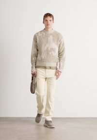 Young man walking, wearing beige textured sweater, cream pants, gray sneakers, and holding a small taupe bag against a plain white background.