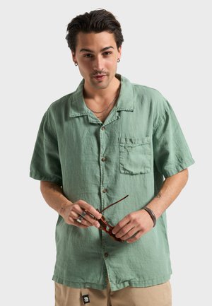 Chemise à manches courtes avec boutons, en lin vert menthe doux, dotée d'une poche poitrine. Le mannequin tient des lunettes de soleil marron et porte des bijoux argentés.