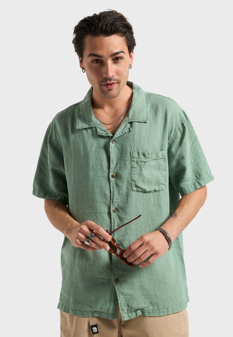 Chemise à manches courtes avec boutons, en lin vert menthe doux, dotée d'une poche poitrine. Le mannequin tient des lunettes de soleil marron et porte des bijoux argentés.