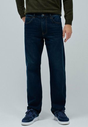Jeans de mezclilla azul oscuro con un diseño de pierna recta, ligero desgaste y costuras en contraste. Combinados con zapatillas de deporte en azul marino.