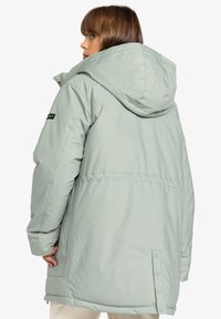 Jachetă puffer cu glugă de culoare verde deschis, cu o croială lejeră, având capse la tiv și un material neted, texturat. Logo-ul Roxy pe mânecă.