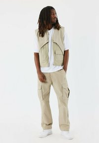 Pantalones cargo beige y un chaleco sin mangas a juego sobre una camiseta blanca. El chaleco tiene bolsillos y una textura suave. Zapatillas blancas completan el conjunto.