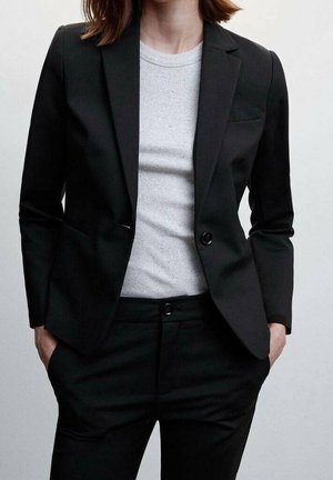 Vrouw met een zwart colbert en broek, een wit geribd shirt met ronde hals, handen in de zakken, tegen een effen lichte achtergrond.