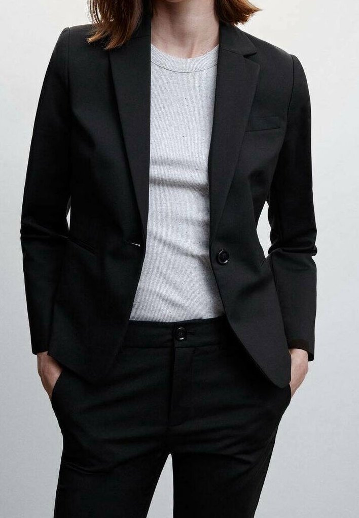 Femme portant un blazer noir et un pantalon avec un t-shirt blanc côtelé à col rond, mains dans les poches, sur un fond clair uni.