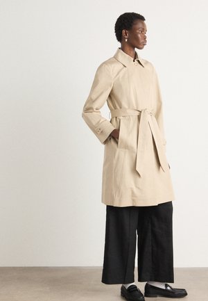 Beige Trenchcoat mit Gürtel, großen Revers und seitlichen Taschen. Über weiten schwarzen Hosen und Slippern getragen. Glatte Textur, minimalistisches Design.