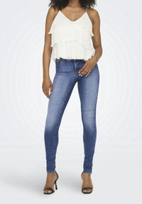 Weißes, ärmelloses Oberteil mit geschichteten Rüschen, kombiniert mit blauen Skinny-Jeans. Das Model trägt schwarze, römische Sandalen mit Absatz. Heller Hintergrund.