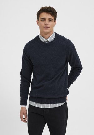 Pulover tricotat bleumarin, cu manșete și tivuri curbate, purtat peste o cămașă cu dungi deschise. Modelul stă în fața unui fundal simplu.