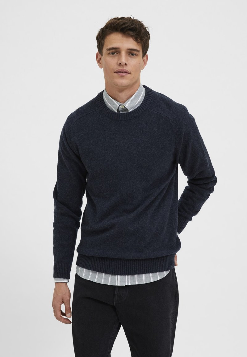 Suéter de punto azul marino con puños y dobladillo acanalados, llevado sobre una camisa de rayas clara. El modelo está de pie frente a un fondo liso.