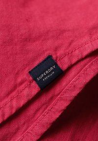 Rode stof met een gestructureerde afwerking en een zwart label. Het label heeft de tekst "SUPERDRY PREMIUM" in witte letters, genaaid aan de rand van de stof.
