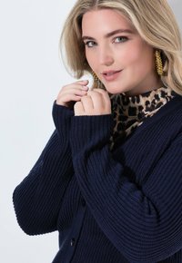 Blond kvinna med gröna ögon, iförd stora guldörhängen, leopardmönstrad topp och marinblå stickad kofta, som håller koftans krage nära sitt ansikte.