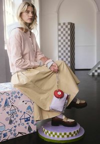 Mladá žena sediaca na dekoračnom bloku, oblečená v ružovej pruhovanej košeli so zapínaním na gombíky, béžovej sukni a hnedých platformových topánkach, držiaca červeno-fialovú tašku.