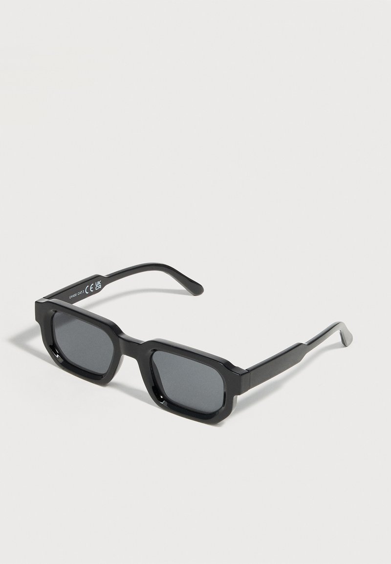 Lunettes de soleil rectangulaires noires avec des montures épaisses et des verres teintés foncés, inclinées sur un fond blanc.