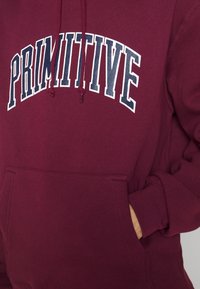 Sweat-shirt bordeaux avec texte "PRIMITIVE" en blanc et marine, cordons de serrage, et une poche avant avec une main partiellement insérée.