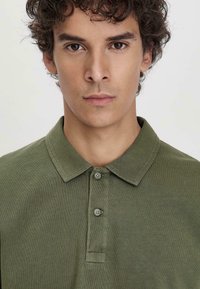 Olijfgroene polo met een kraag, twee knopen en een zachte, gestructureerde stof. Eenvoudig ontwerp zonder patronen of accenten.