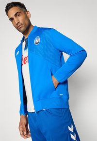 Blå atletisk jakke med mesh-paneler, med et logo på brystet og lynlåslukning. Tekstureret stof og en afslappet pasform.